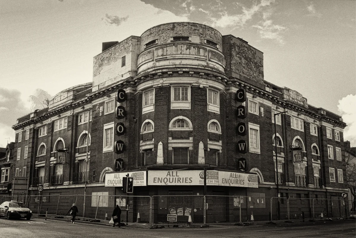 Sepia-toned photograph of the derelict Crown cinema building on a street corner, its vertical CROWN signage still in place above boarded windows and ALL ENQUIRIES hoardings