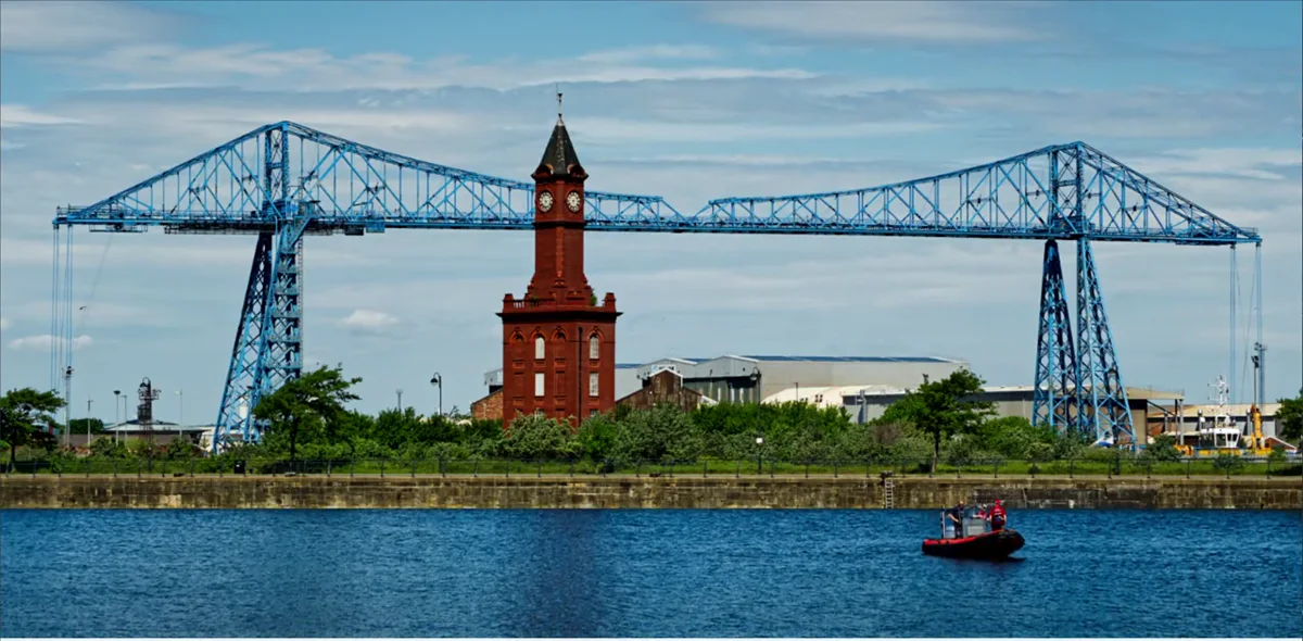 The Middlesbrough Transporter Bridge in blue steel spanning the River Tees, with the red brick clock tower of the old customs house centred beneath it and a small red boat on the water